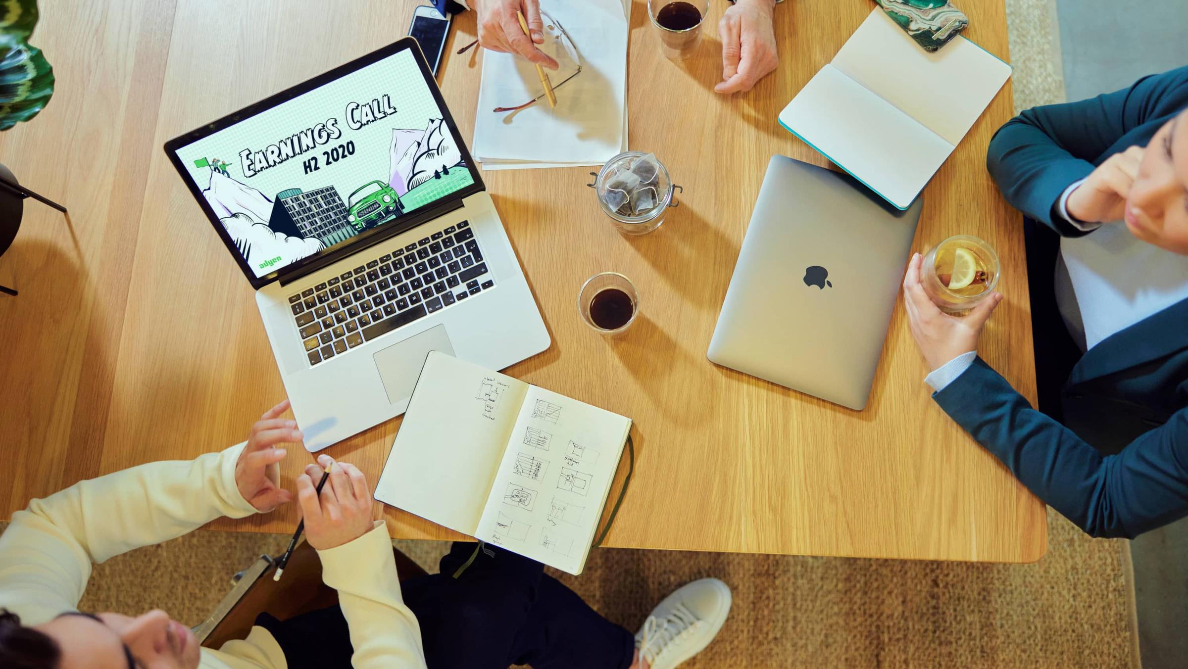Top view of a work meeting with laptops, notepads, and coffee on a wooden table with an Adyen Earnings Call on laptop screen.