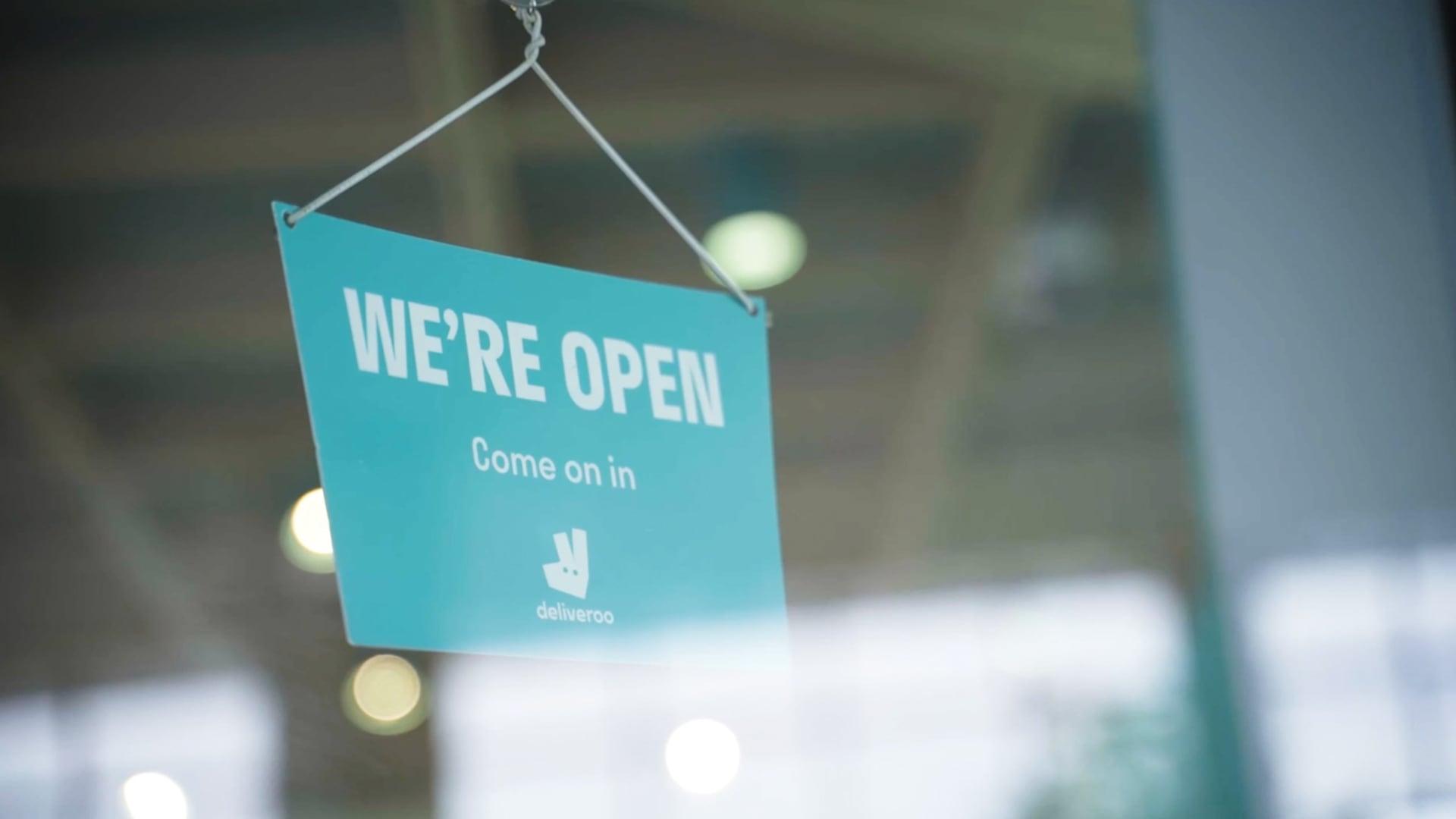 A teal 'WE'RE OPEN' sign by Deliveroo hanging on a glass door.