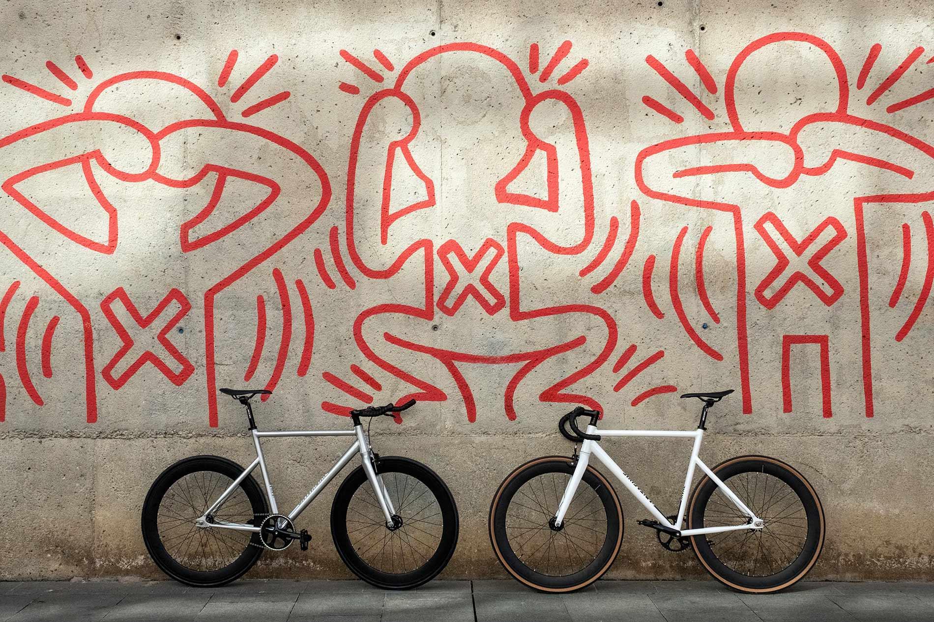 Dos bicicletas aparcadas frente a una pared con graffiti rojo de figuras de ciclismo estilizadas.