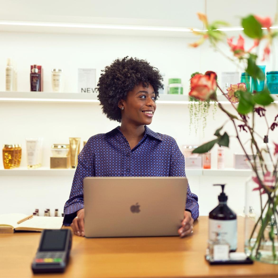 Smiling woman sitting at a retail checkout desk with a laptop and payment terminal.