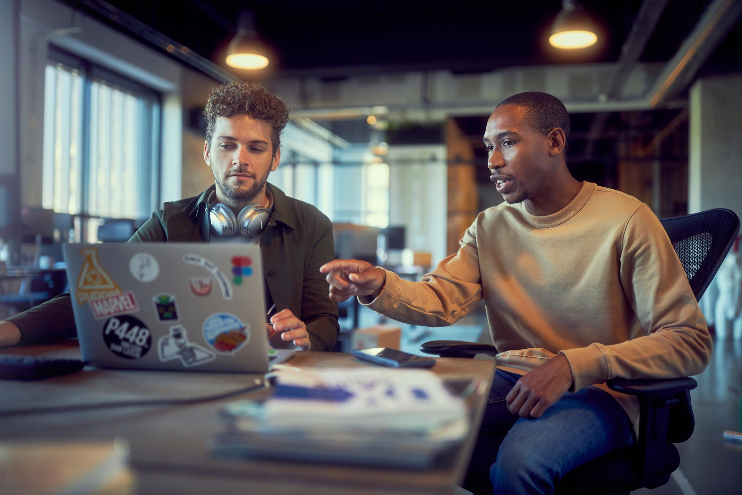 Zwei Kollegen sitzen hinter einem Laptop und diskutieren miteinander in einem Büroumfeld