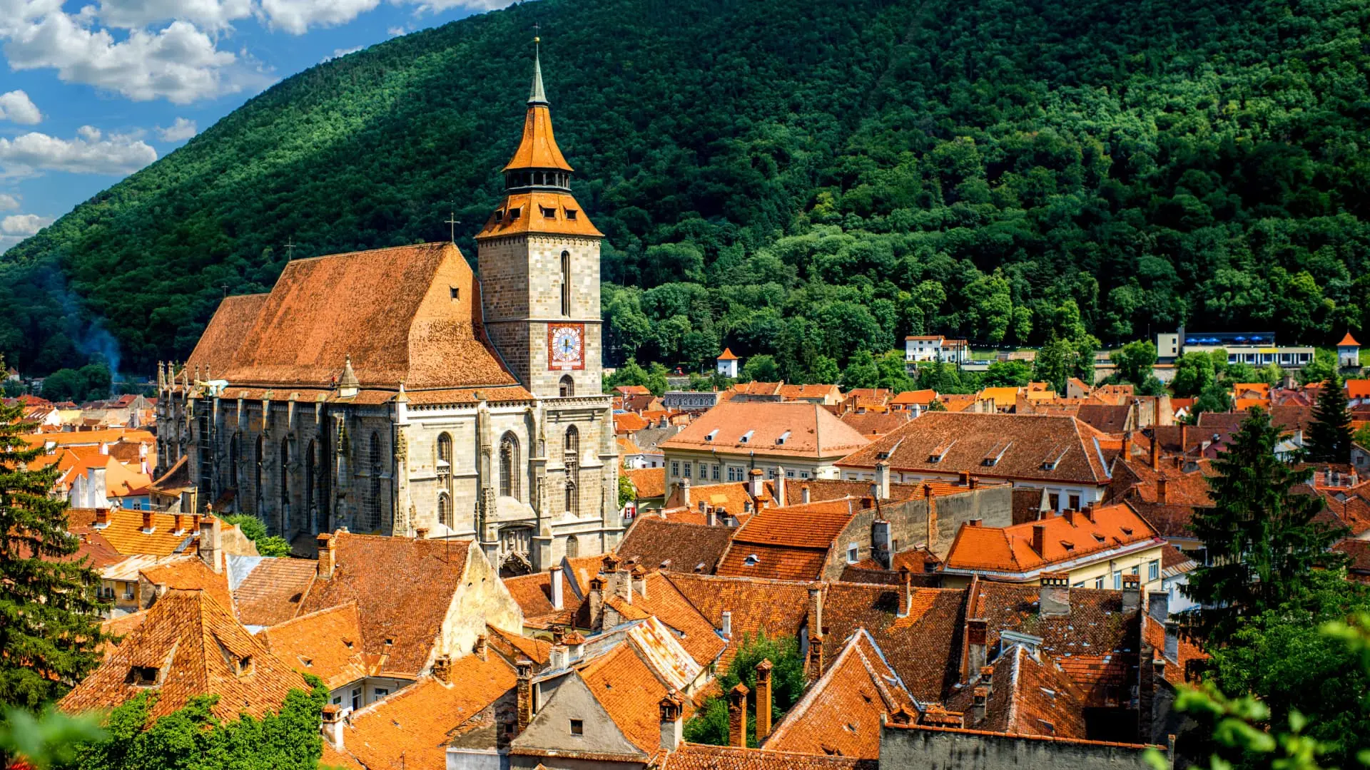 A historic town with orange-tiled roofs and a prominent church tower against a verdant hillside in Romania.