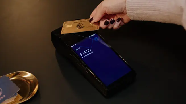 Uma mão segura um cartão American Express enquanto paga pessoalmente em um terminal de pagamento.