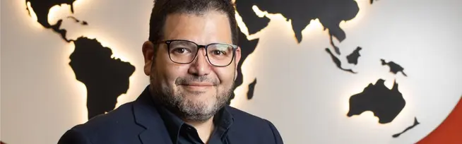 Homem de óculos e barba sorrindo em frente a um mapa-múndi