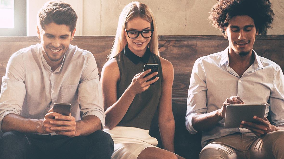 Tres adultos jóvenes utilizando teléfonos inteligentes y una tableta, sentados en un banco en interiores.