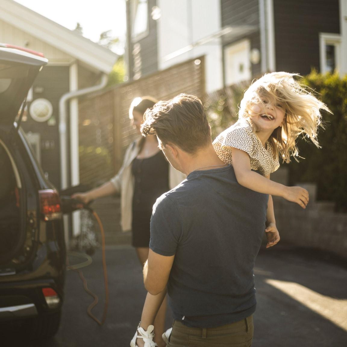Mann løfter et lykkelig barn i luften ved siden av en bil med en kvinne i bakgrunnen