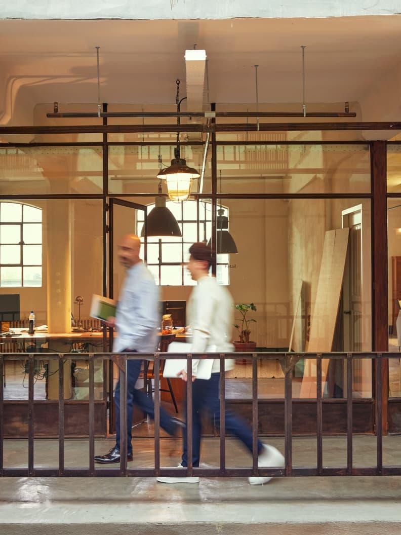 Two people in motion walking through a modern office with large windows and hanging lights.