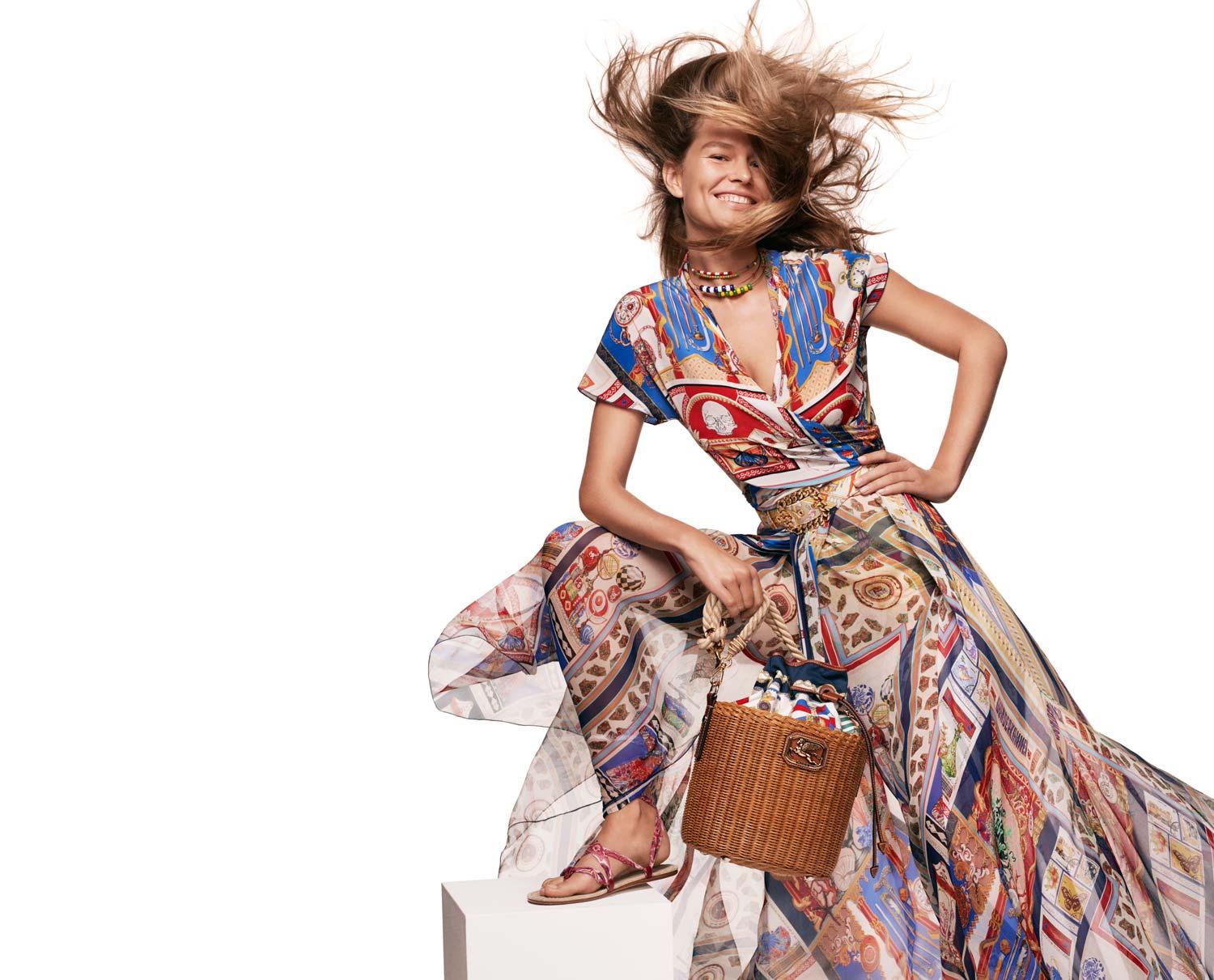 Donna in un abito a fantasia vibrante con i capelli al vento, che tiene una borsa di vimini.