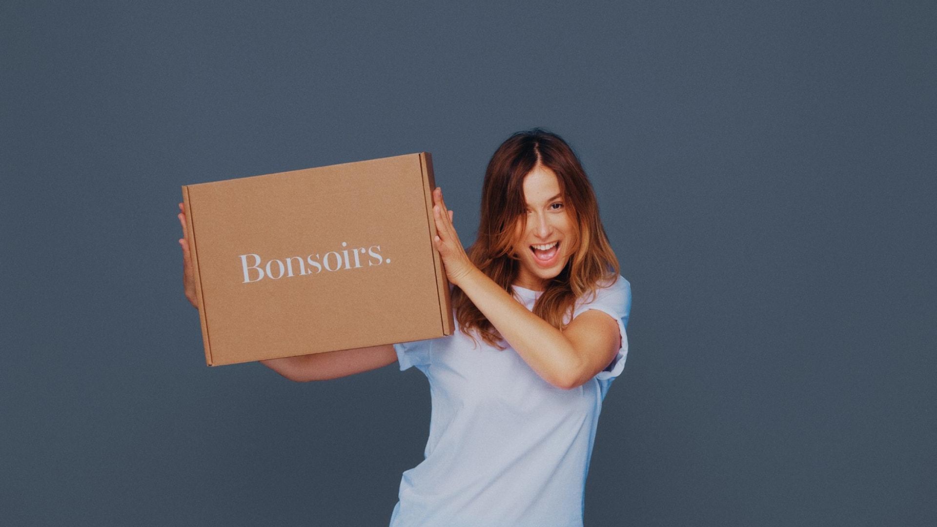 Femme tenant avec joie une boîte en carton avec 'Bonsoirs' écrit dessus.