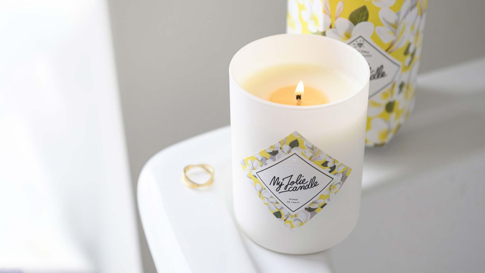 Une bougie parfumée allumée avec une bague à côté sur une surface blanche.