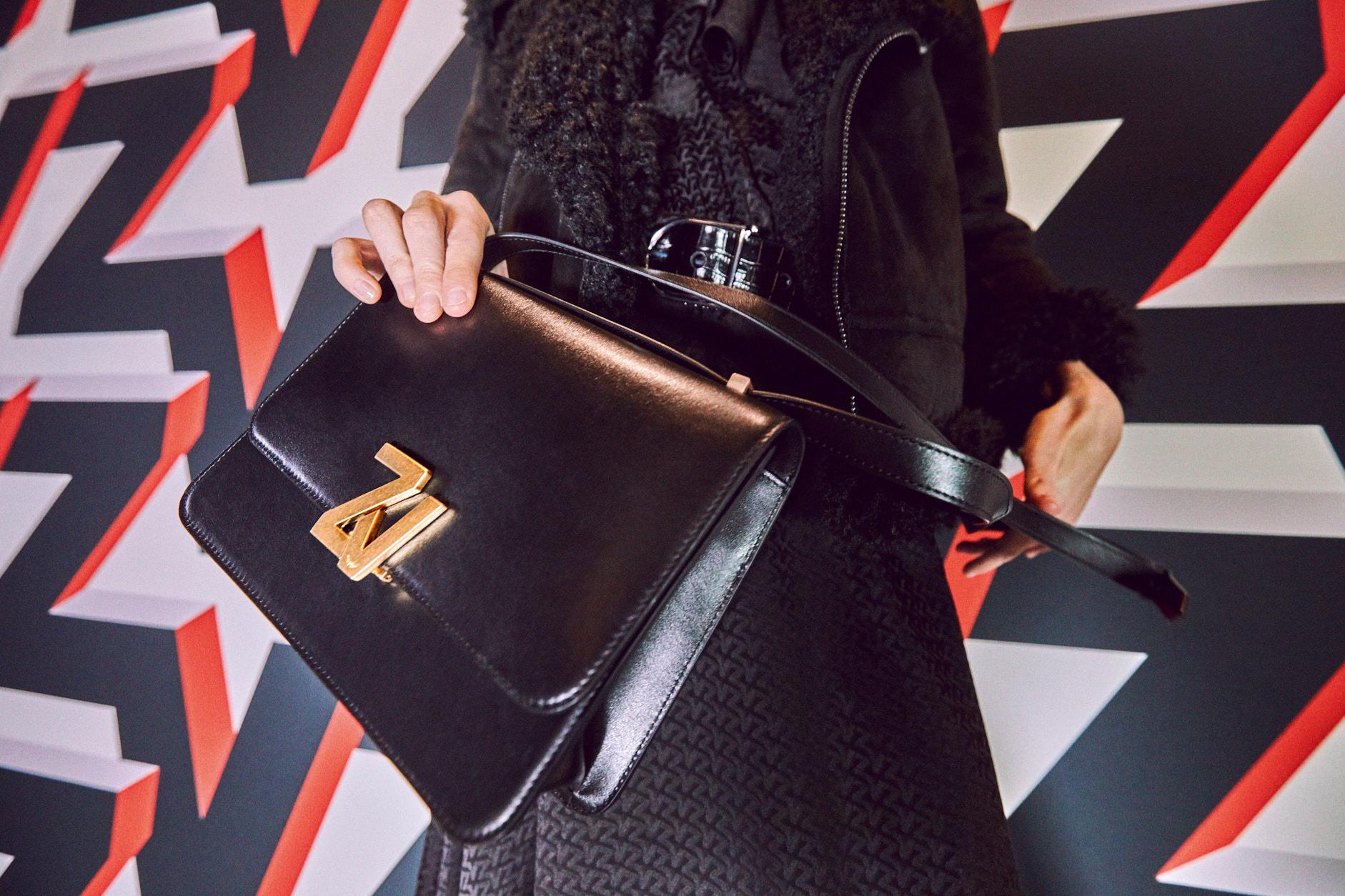 Close-up of a stylish black leather bag with a golden logo, held by a person in a textured coat.