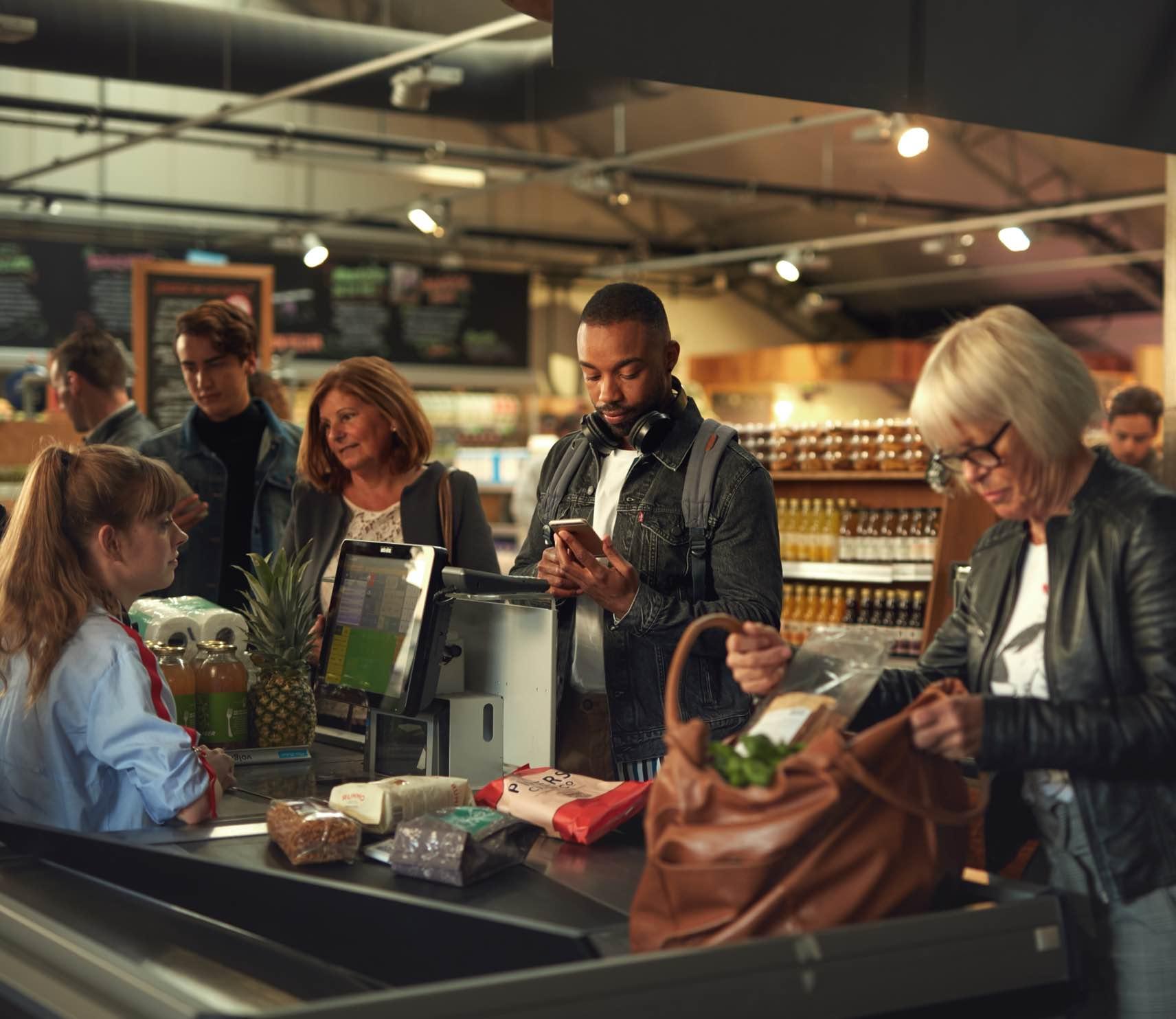 Clientes no caixa de um supermercado, um usando um smartphone possivelmente para pagamento