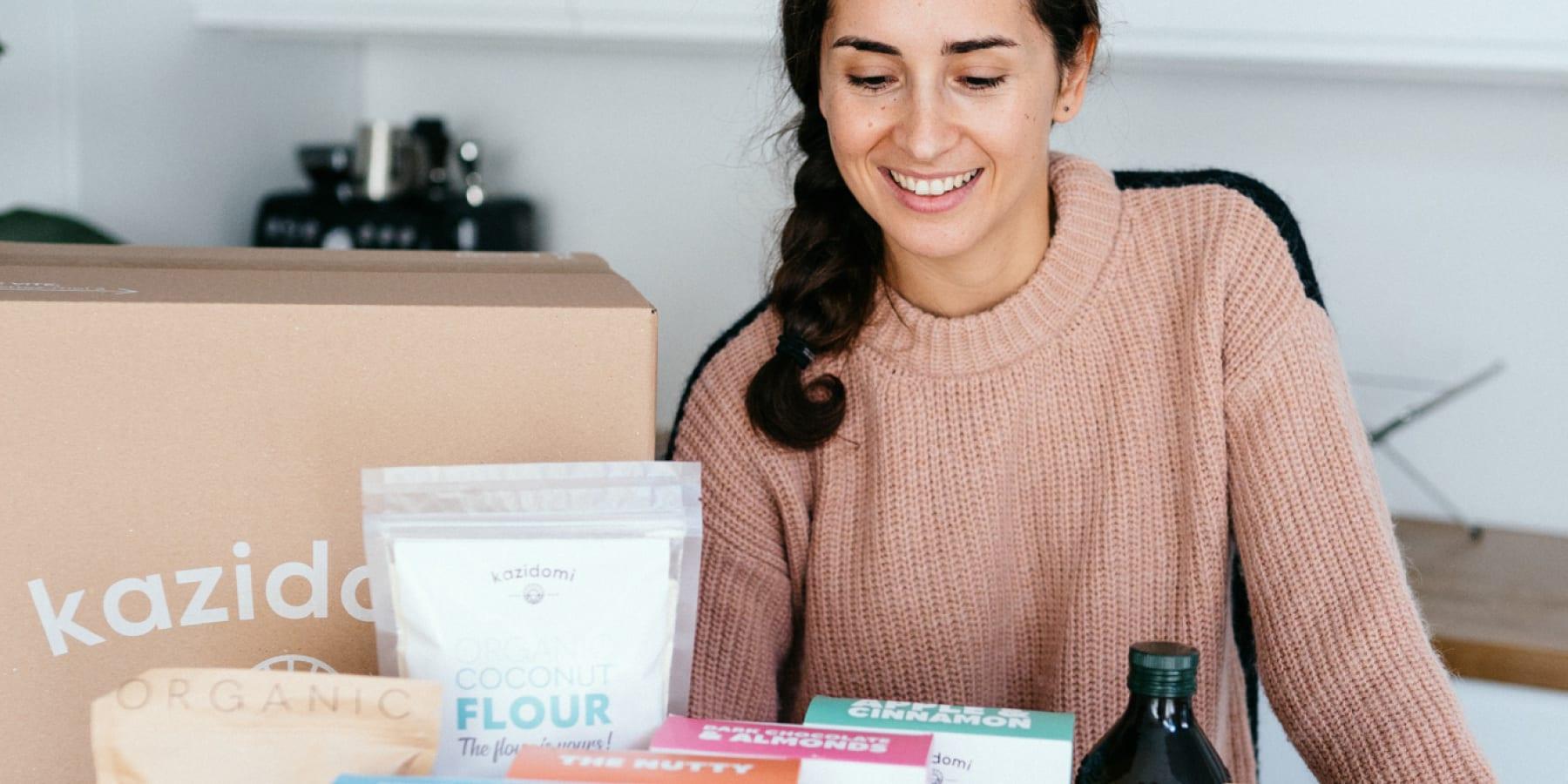 Femme souriante déballant une boîte de produits alimentaires biologiques sur un comptoir de cuisine.