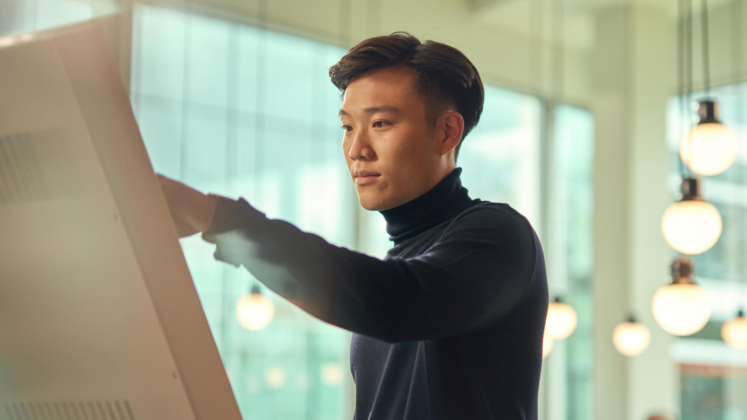 Mann im Rollkragenpullover interagiert mit einem Touchscreen-Display in einem modernen Büroumfeld.