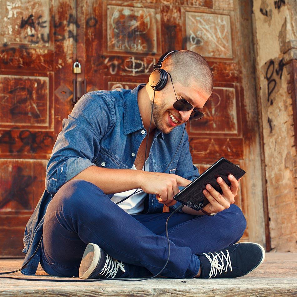 Smilende mann med solbriller og hodetelefoner som bruker en nettbrett mens han sitter på bakken.