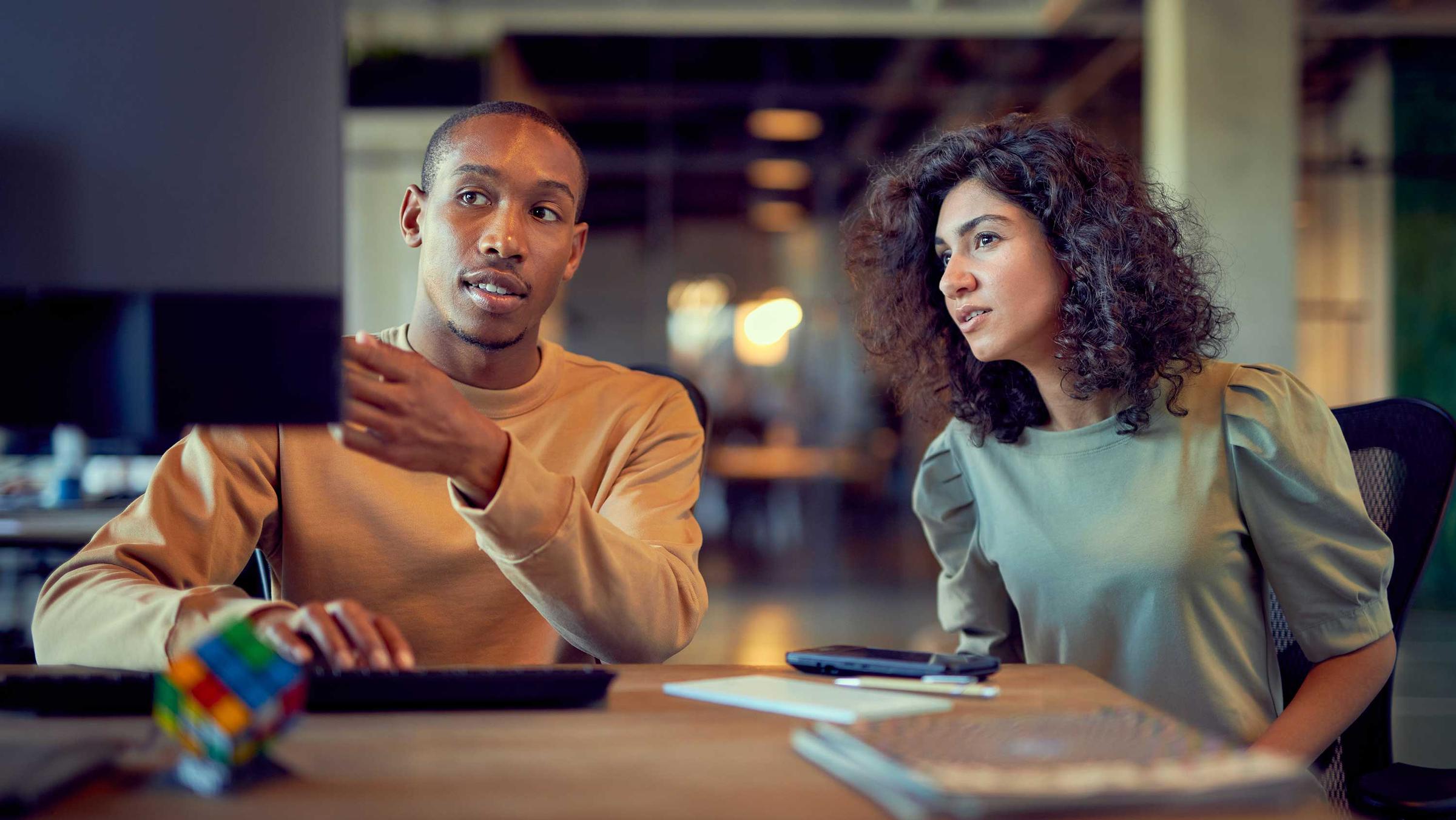 Two professionals discussing work with a computer and documents on desk.