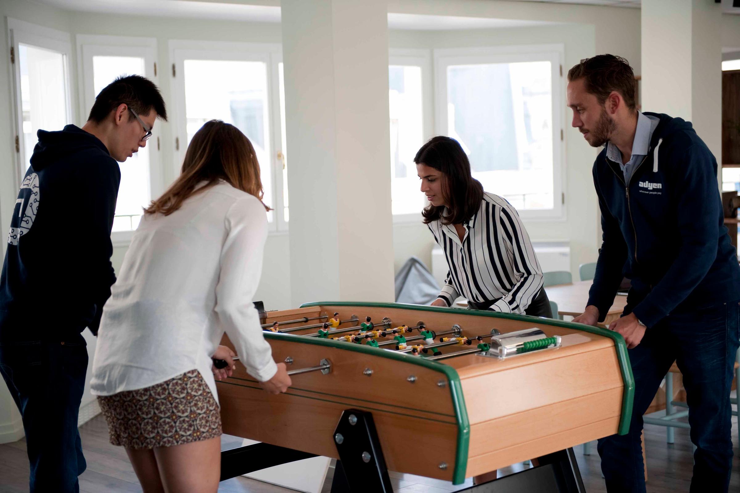 Três pessoas jogando futebol de mesa no escritório, uma usando uma blusa com o logotipo da Adyen