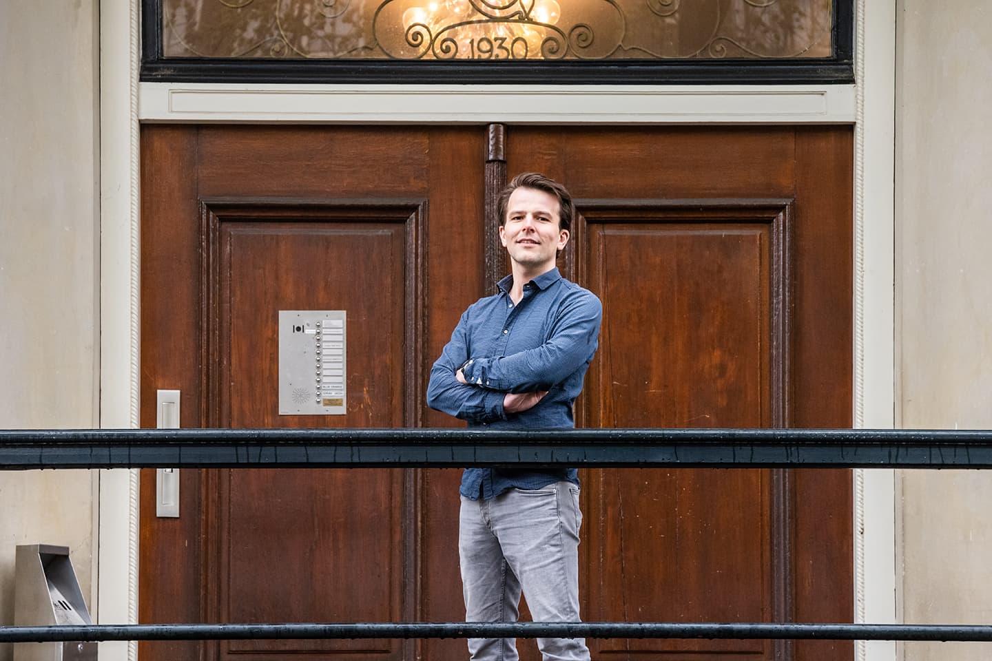 Man staat zelfverzekerd met zijn armen over elkaar voor een houten deur.