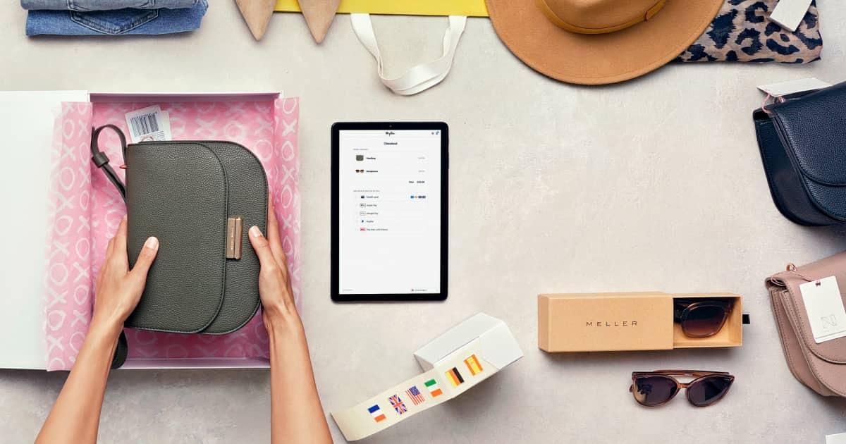 Nadhled osoby balící si tašku s tabletem zobrazujícím platební rozhraní Adyen vedle módních doplňků.