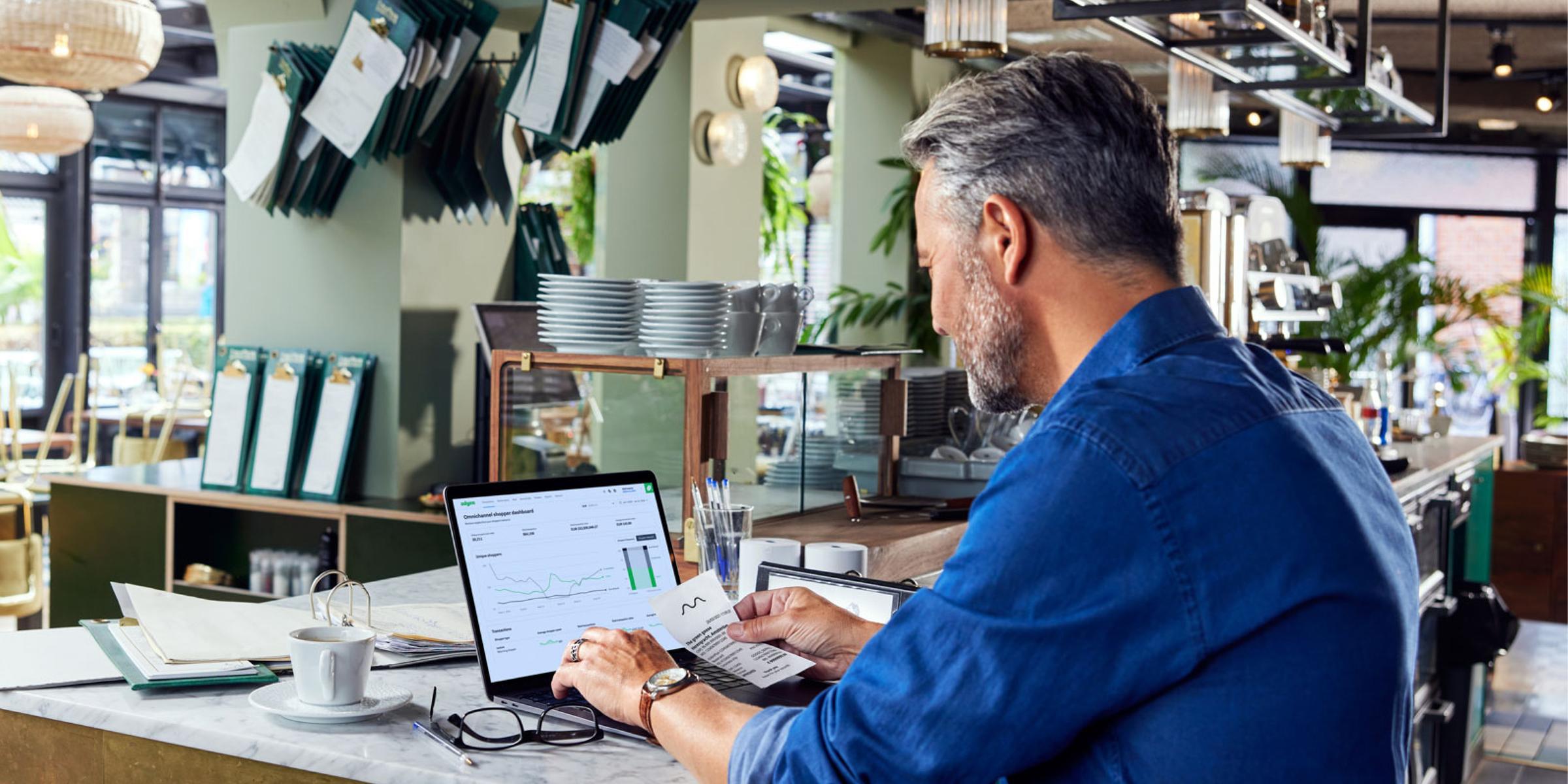 Man analyzing Adyen sales data on laptop in a cafe with bill and cup of coffee on table.