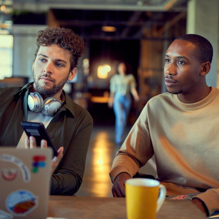 Dois homens sentados a uma mesa, um com fones de ouvido ao pescoço olhando para um smartphone.