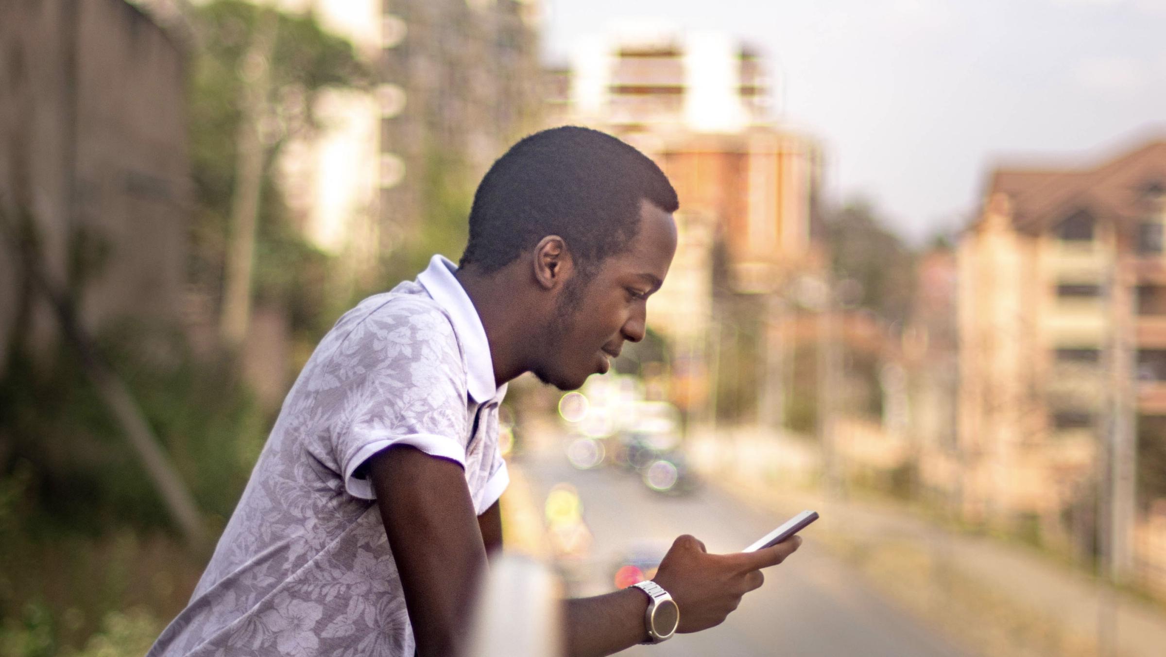 Man in white patterned shirt looking at smartphone by the roadside.