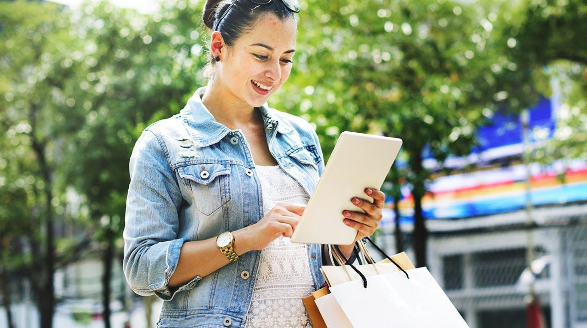 Mujer feliz utilizando una tableta mientras lleva bolsas de compras en una calle soleada.