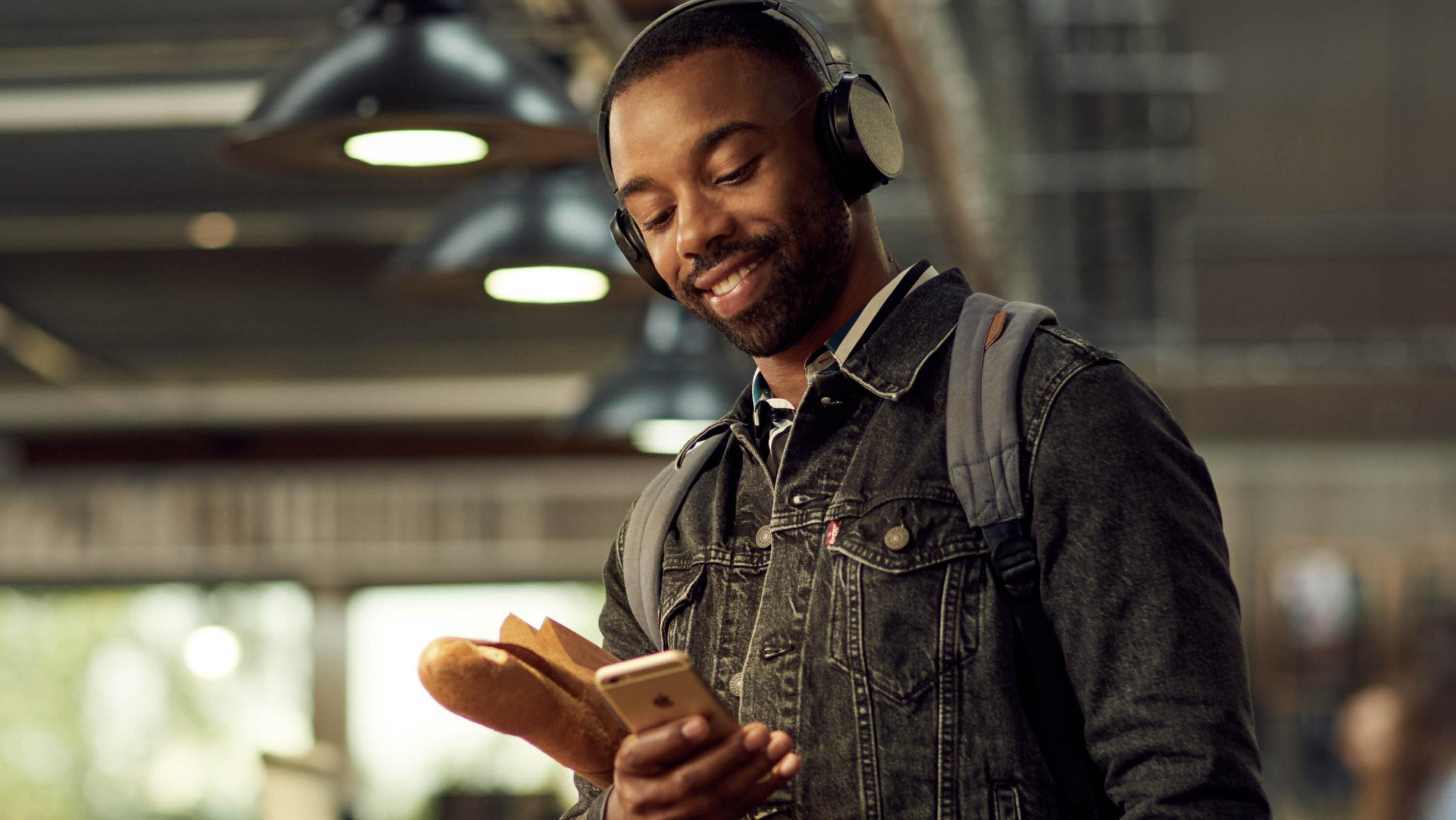Smiling man wearing headphones using smartphone while holding a baguette.