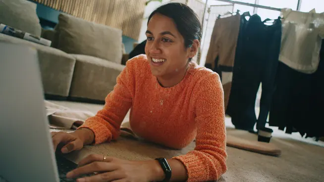 Mulher sorrindo deitada no chão e usando laptop em um quarto aconchegante com cabide na parte traseira.
