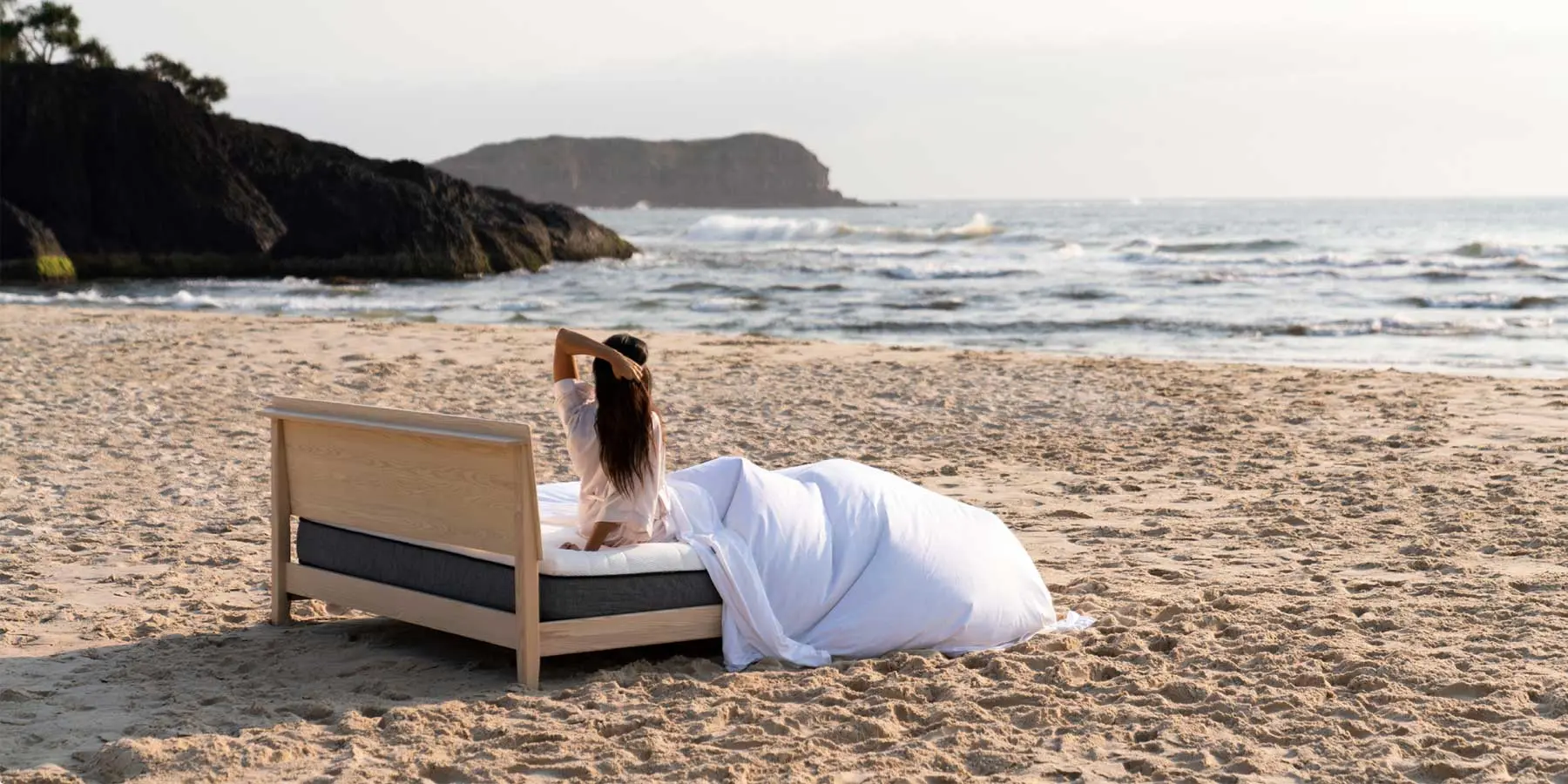 Person relaxing on a bed with white sheets on a sandy beach, facing the ocean.
