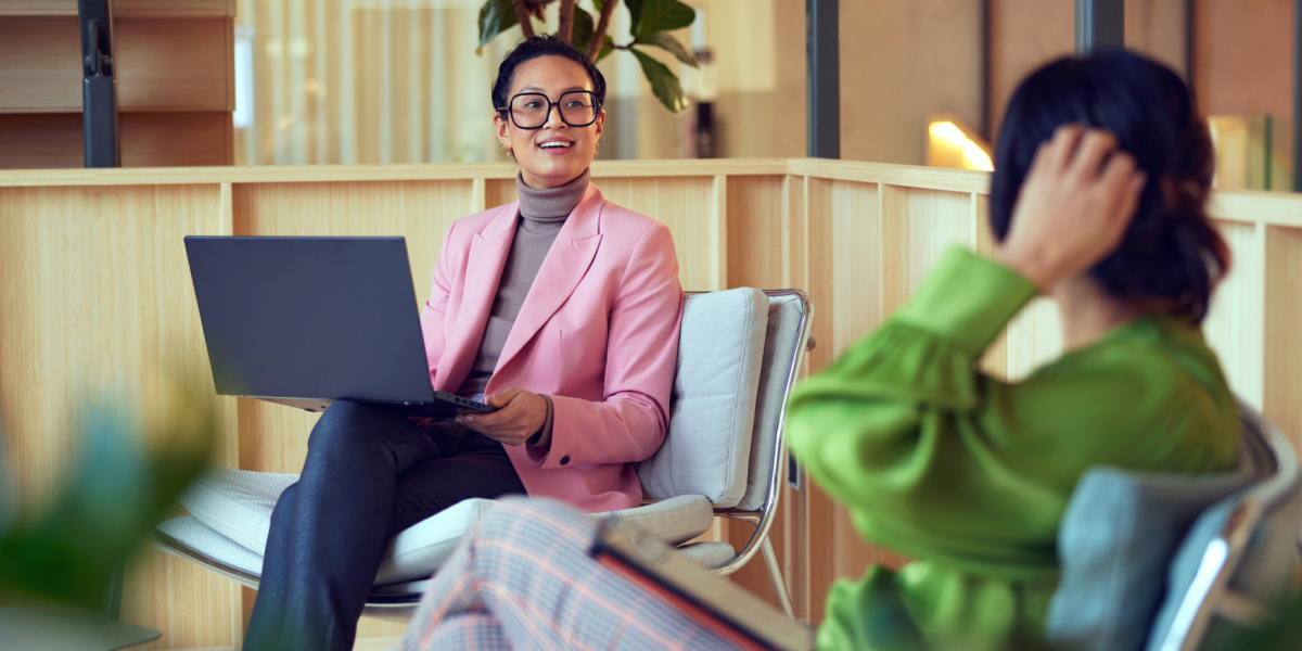 Duas mulheres profissionais em um ambiente de escritório informal com uma usando um laptop.