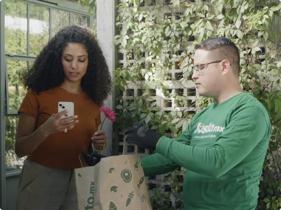 Persona de entrega con camisa verde entregando una bolsa de comestibles a una mujer que sostiene un smartphone.