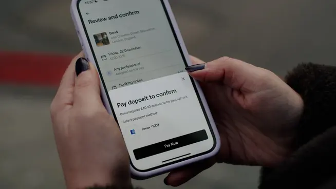 Dos manos sosteniendo un teléfono y pagando un corte de pelo en la aplicación de reserva Fresha con American Express.