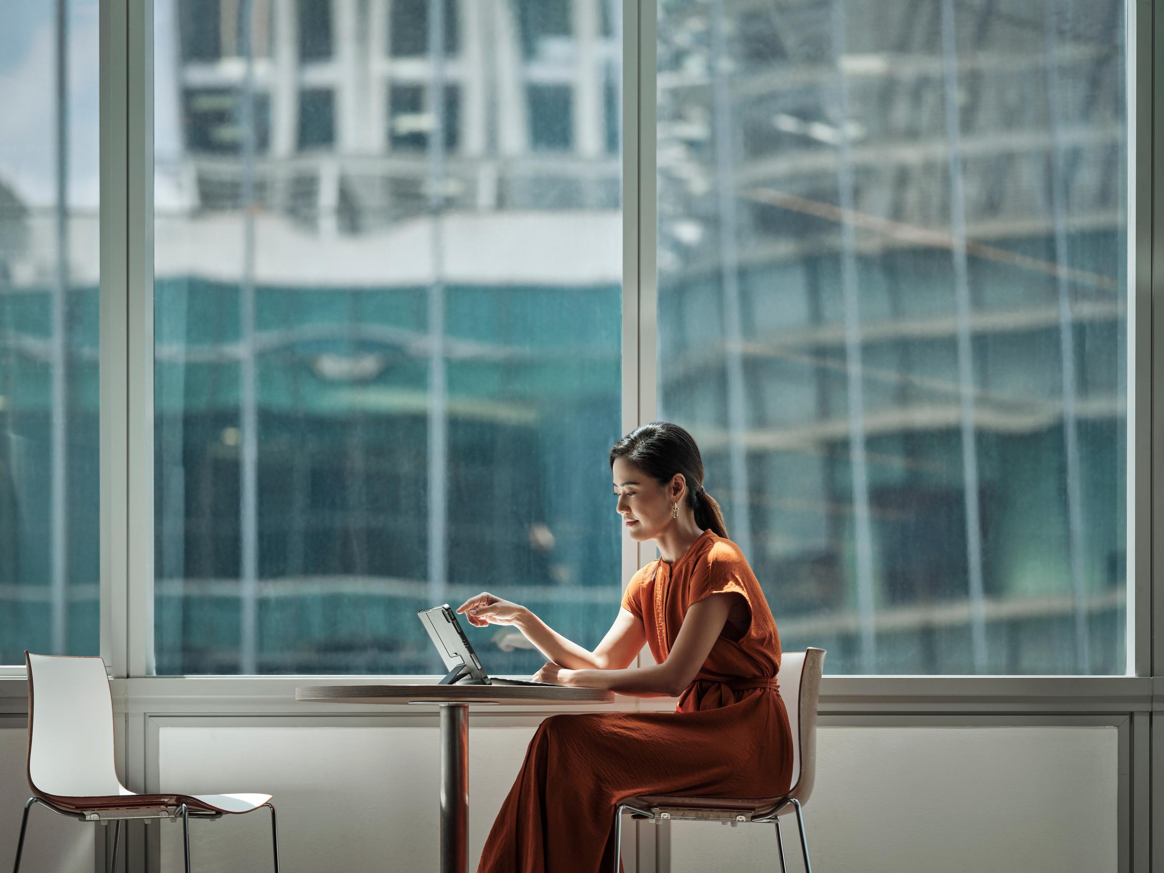Une femme assise à une table à côté d'une grande fenêtre regardant son ipad