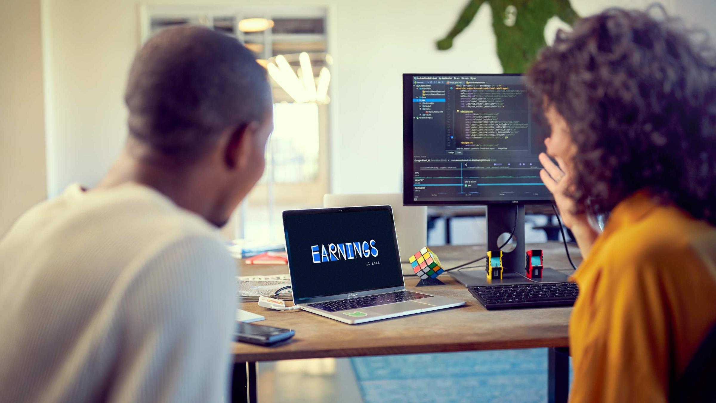Two people collaborating in an office with multiple computer screens displaying code and financial data.