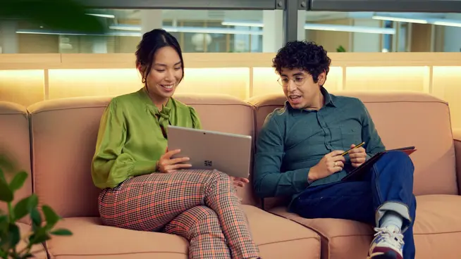 Two individuals seated on a couch, one holding a tablet and the other taking notes.