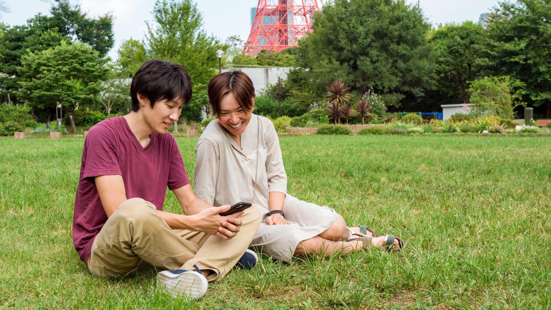 Two people sitting on the grass, looking at a phone, and smiling