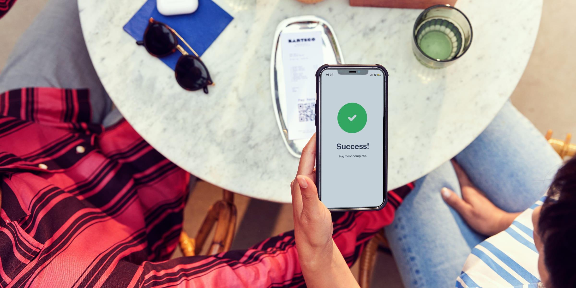 Persona que sostiene un smartphone con un mensaje de éxito en la pantalla después de completar un pago en un café al aire libre.