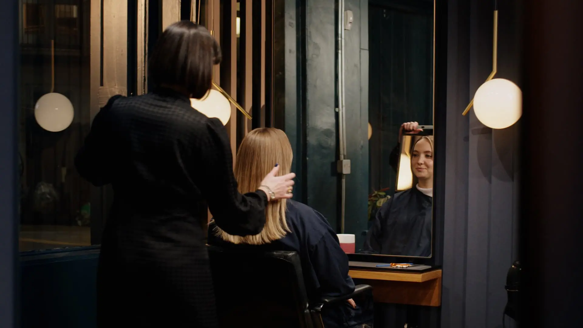 Une femme est assise dans un salon en train de regarder sa nouvelle coupe de cheveux avec le coiffeur assis derrière le fauteuil.