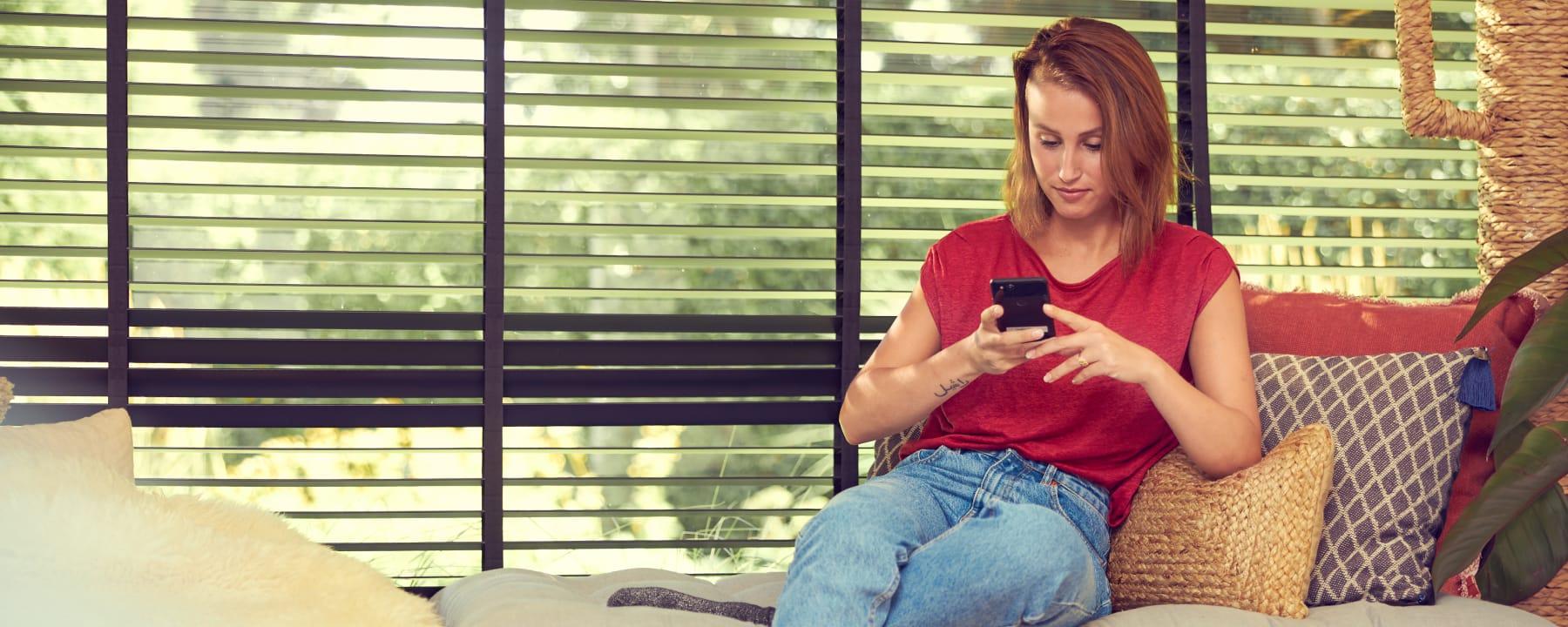 Mujer sentada en un sofá utilizando un smartphone con persianas en el fondo.