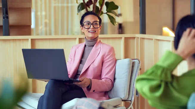 Smiling woman in a pink blazer working on a laptop seated opposite a person in green.