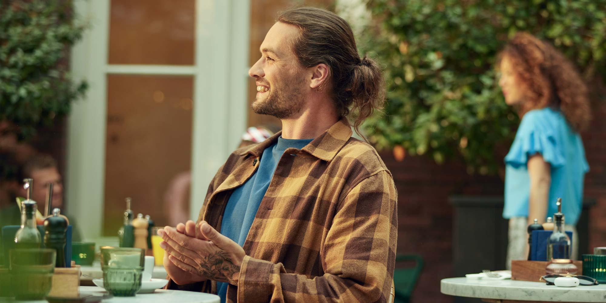Man with a ponytail smiling and clapping his hands at an outdoor cafe table.