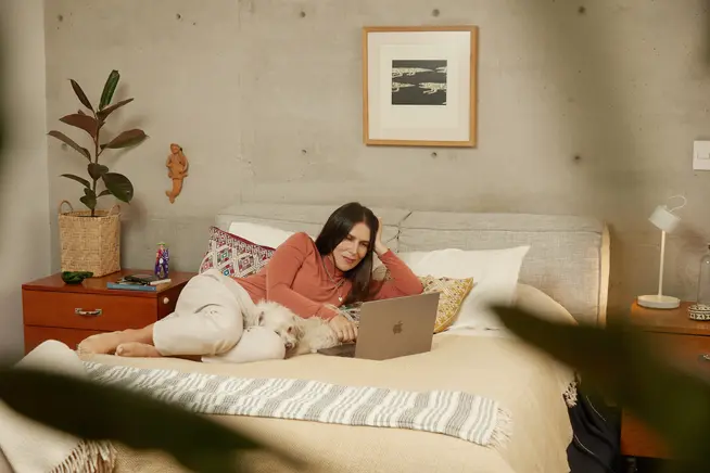 Kobieta leżąca na łóżku ze swoim psem, używająca laptopa.