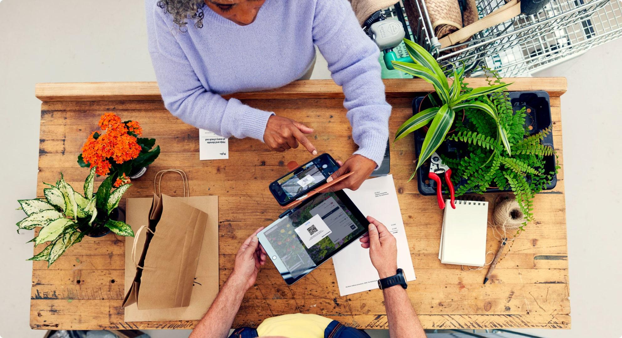 Top view of two people at a wooden table using a smartphone and a tablet to make a contactless payment with plants and documents.