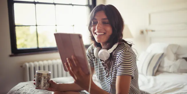 Smiling woman with headphones around her neck holding a tablet and a cup of coffee.