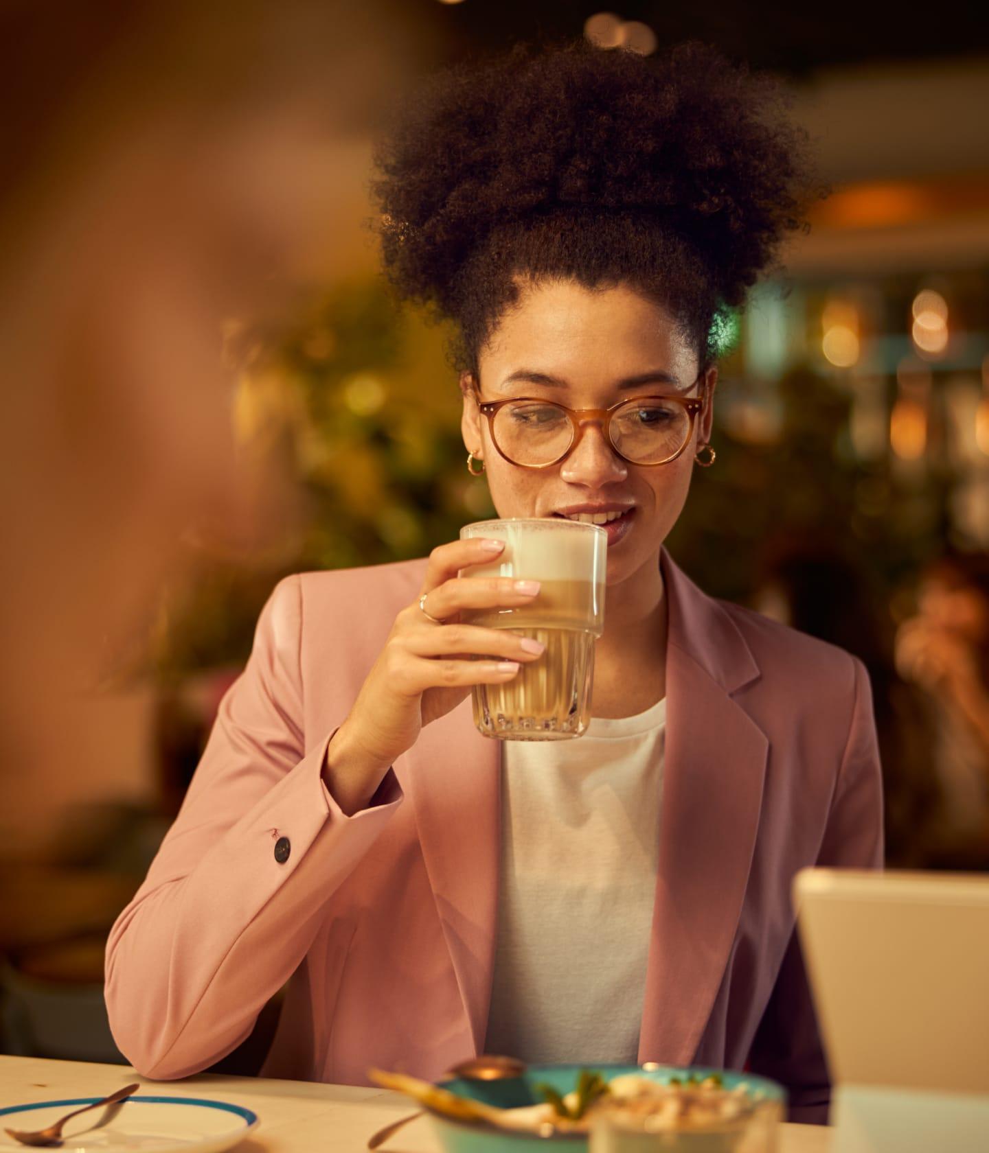Woman in pink blazer drinking a glass of latte at a cafe with a laptop on the table.