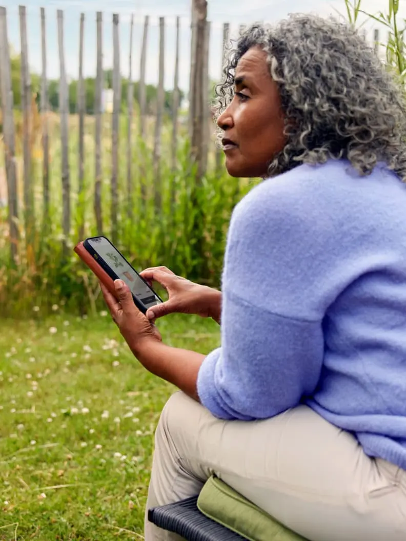 A woman sitting outdoors holding a smartphone with a focus on the screen.