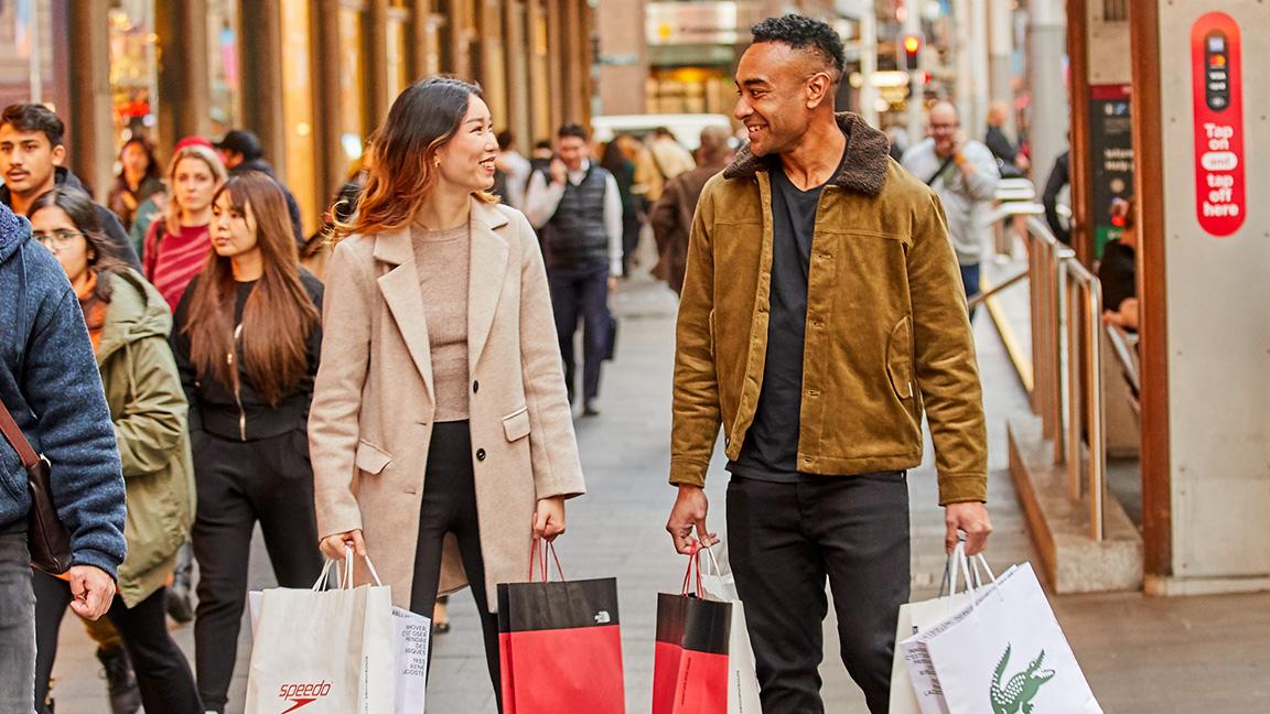 Two people happily walking and carrying shopping bags in a busy urban street.