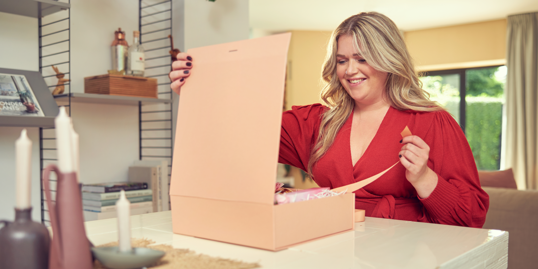 Smiling woman in a red dress opening a large pink gift box at home.