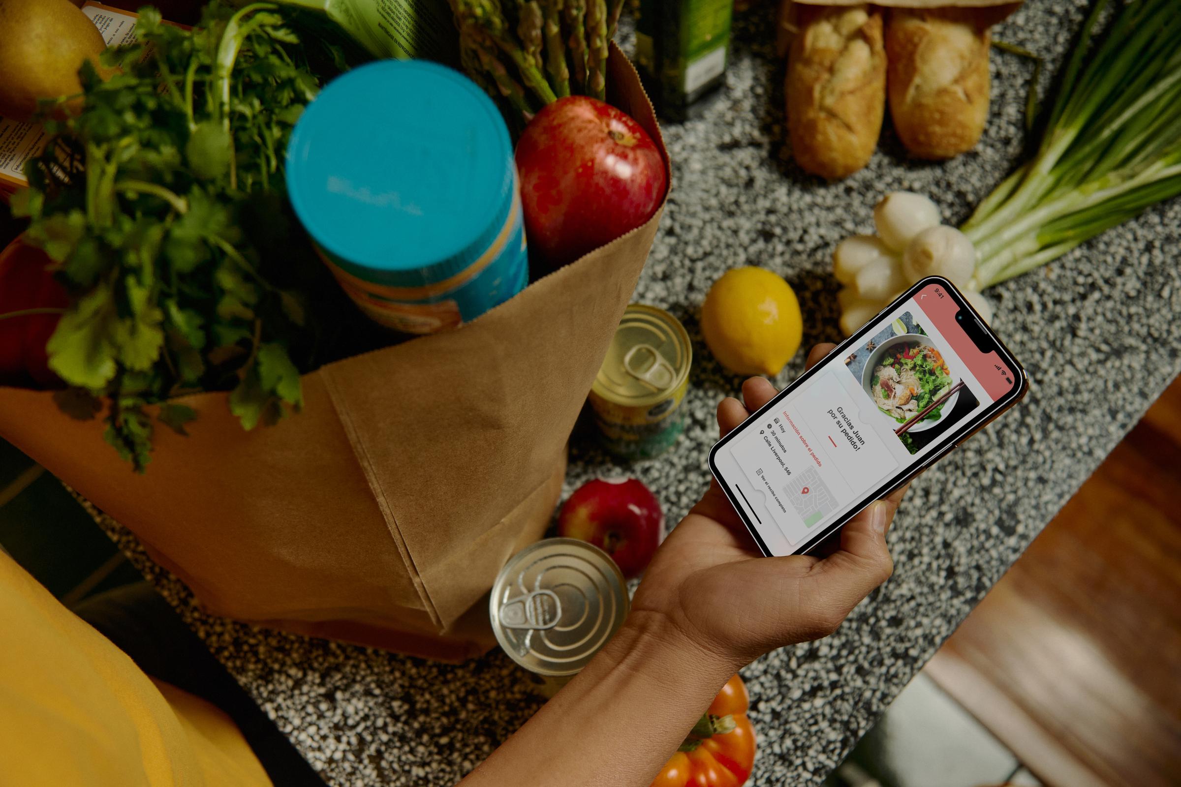 Primo piano di una mano che tiene uno smartphone che visualizza un'app di consegna di cibo, circondata da generi alimentari.